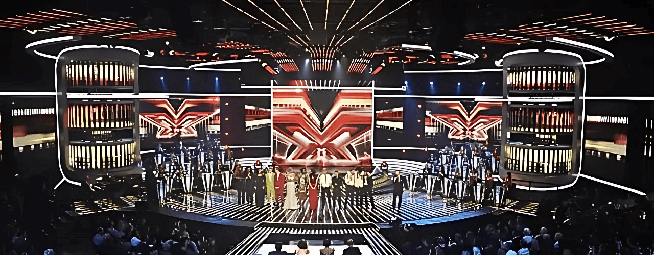 A large stage with bright lights and a panel of judges. Performers stand in a line, with a stylized "X" design in the background. Audience members sit in front.